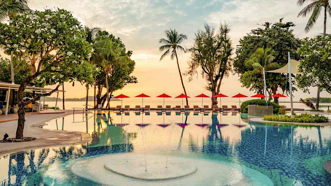 En utomhuspool med en fontän, omgiven av solstolar och röda parasoller. I bakgrunden syns träd och en stilla kustlinje vid solnedgång. (AI-genererad text)