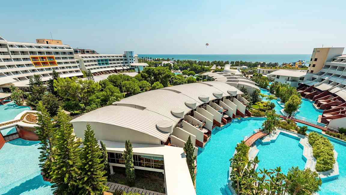 Vy över ett hotellområde med moderna byggnader, stora pooler och prunkande trädgårdar som sträcker sig ut mot havet. (AI-genererad text)