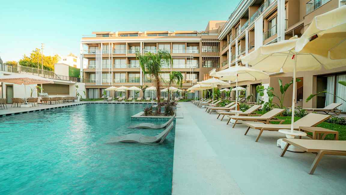 En hotellpool med solsängar och parasoller bredvid moderna byggnader. (AI-genererad text)