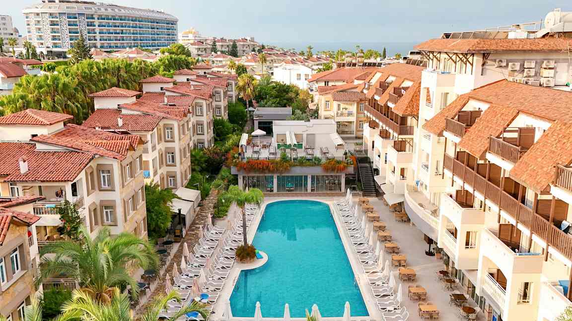 Hotellområde med en pool omgiven av solstolar och byggnader med röda tak. (AI-genererad text)