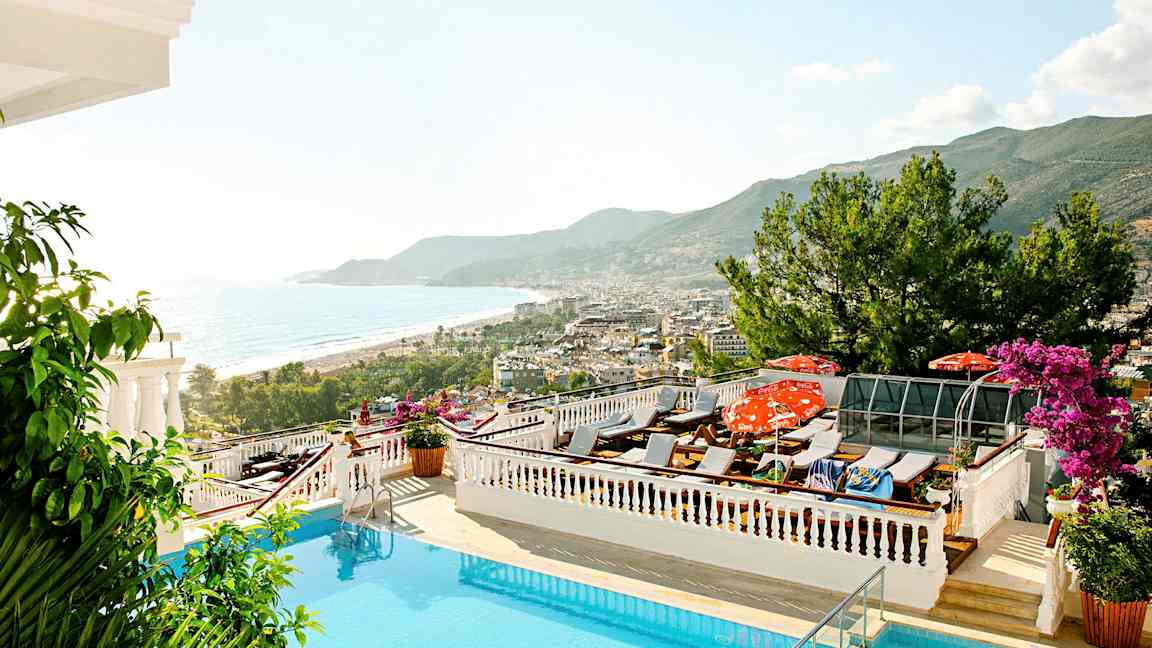 Utsikt över en pool med solstolar, havet och en kuststad i bakgrunden. (AI-genererad text)