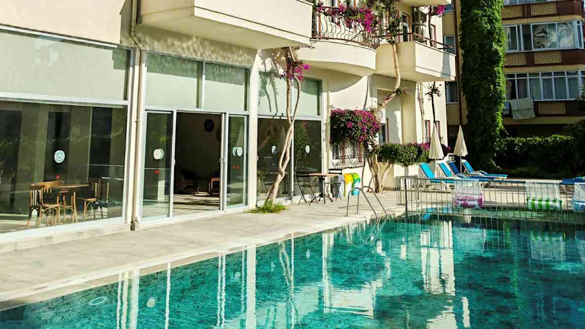 En pool framför en byggnad med balkonger och stora glasfönster. Bilderna visar även stolar och bord samt parasoller vid poolområdet. (AI-genererad text)