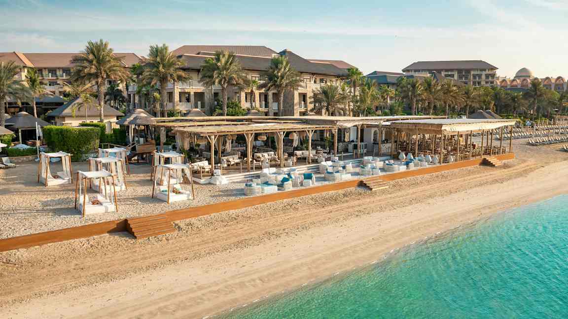 Strand med solstolar, parasoller och ett hotell i bakgrunden. (AI-genererad text)