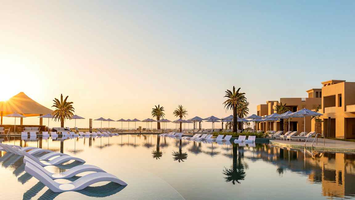 En vy av en infinitypool med solstolar och parasoller, omgiven av palmer och semesterbostäder vid solnedgång. (AI-genererad text)