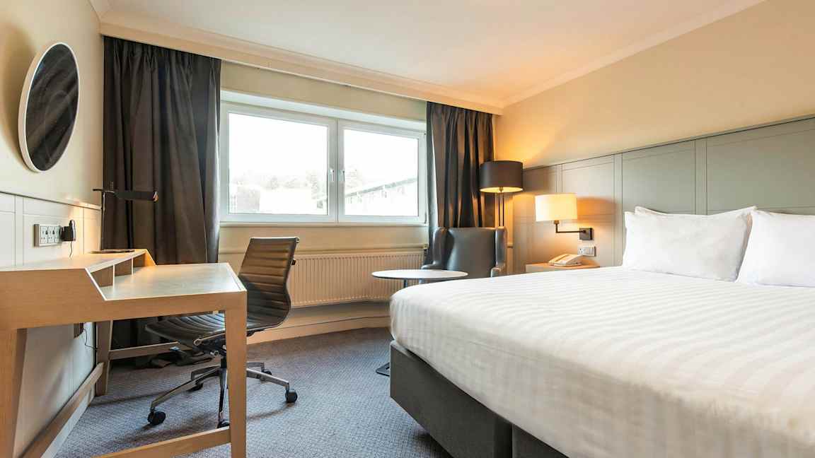 Bild på ett hotellrum med en säng, skrivbord och stol, och en fåtölj bredvid ett fönster. (AI-genererad text)