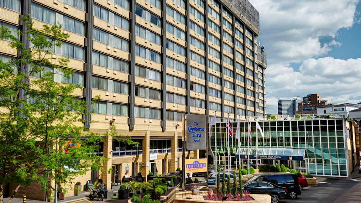 Hotellbyggnad med flera våningar, en glasentré och en parkeringsplats framför. (AI-genererad text)