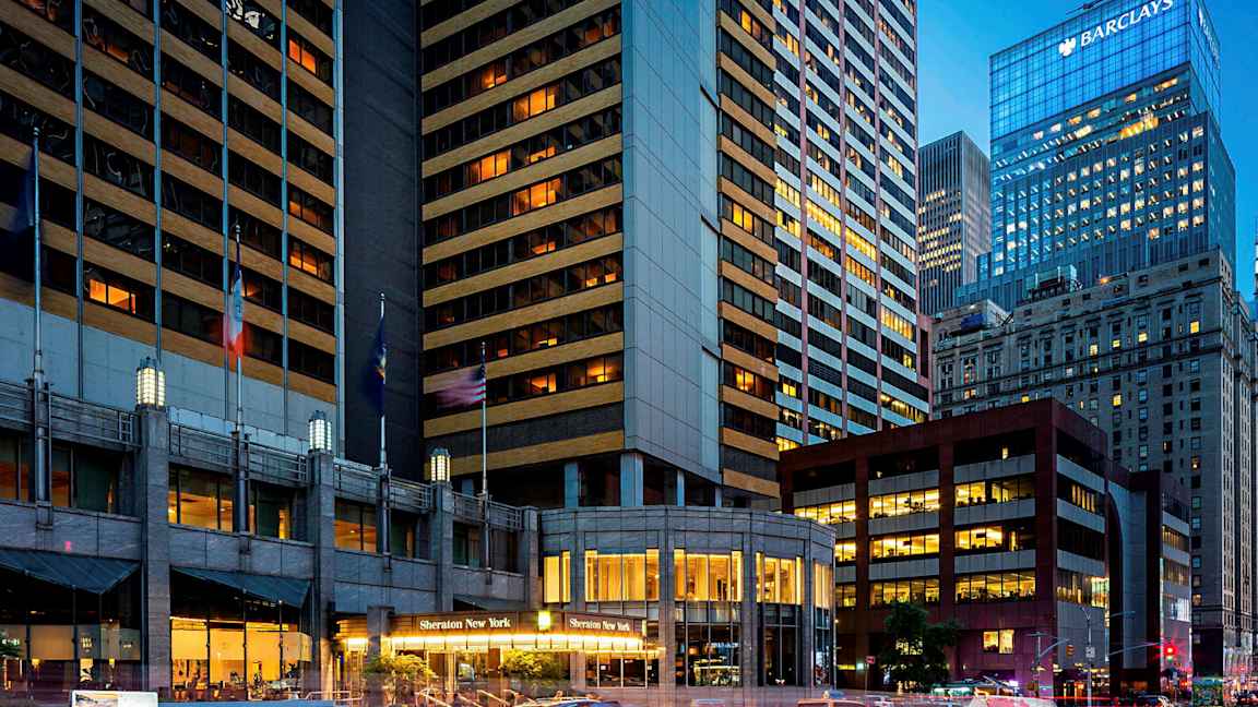 En bild av en stadsmiljö med höga byggnader, varav en byggnad har texten "Sheraton New York" vid dess ingång. Taxibilar syns på gatan. (AI-genererad text)