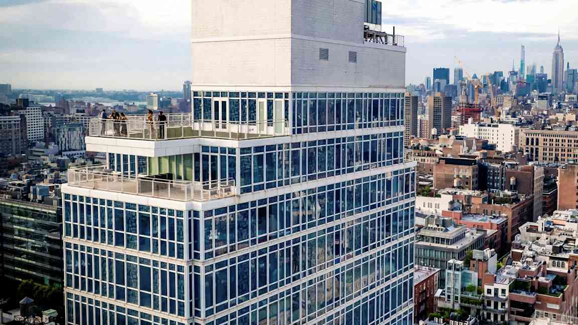 Hög byggnad med glasfasad och terrass, omgiven av stadsmiljö. (AI-genererad text)