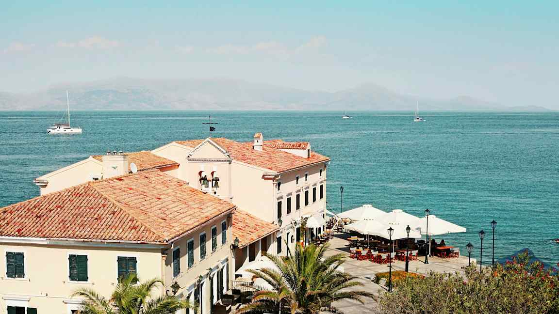 Bild av byggnader med tegeltak vid havet, palms och parasoller synliga. (AI-genererad text)