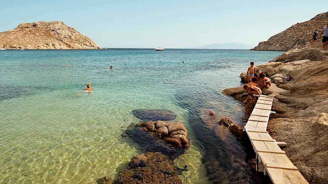 Personer vid en klippa nära en klarblå havsvik med badande. (AI-genererad text)