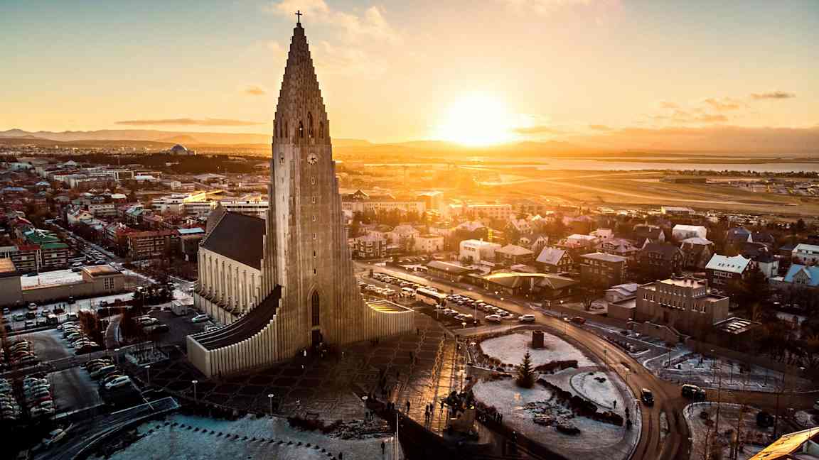 En kyrka med modern arkitektur och en hög tornstruktur, omgiven av en stad och ett landskap under en solnedgång. (AI-genererad text)