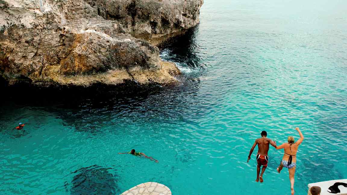 Två personer hoppar från en klippa ner i blått vatten. (AI-genererad text)