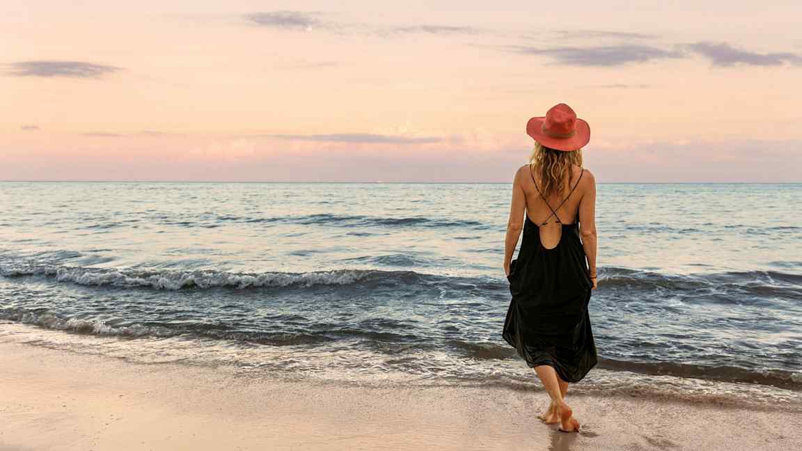 En person med en röd hatt och svart klänning går på en sandstrand vid havet under en pastellfärgad himmel. (AI-genererad text)