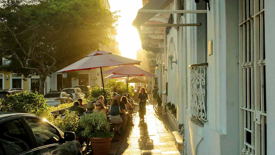 En gata med utomhuscafé där människor sitter under röda parasoller. (AI-genererad text)