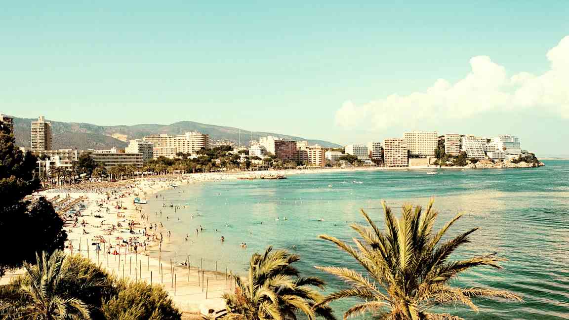 En strand vid havet omgiven av byggnader och palmer. (AI-genererad text)