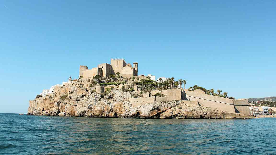 En borg och byggnader på en klippa vid havet, omgivna av palmer. (AI-genererad text)