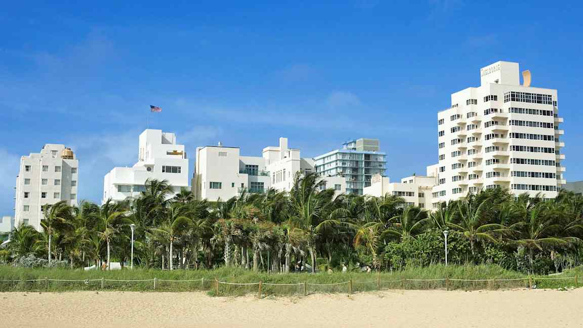 Vita byggnader bakom palmer och sandstrand. (AI-genererad text)