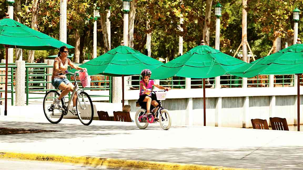 Personer cyklar vid ett område med gröna parasoll och trädkantad bakgrund. (AI-genererad text)