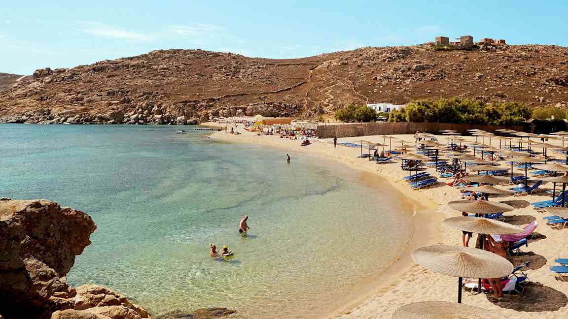 En strand med parasoller och solstolar, folk badar i det klara blåa vattnet. (AI-genererad text)