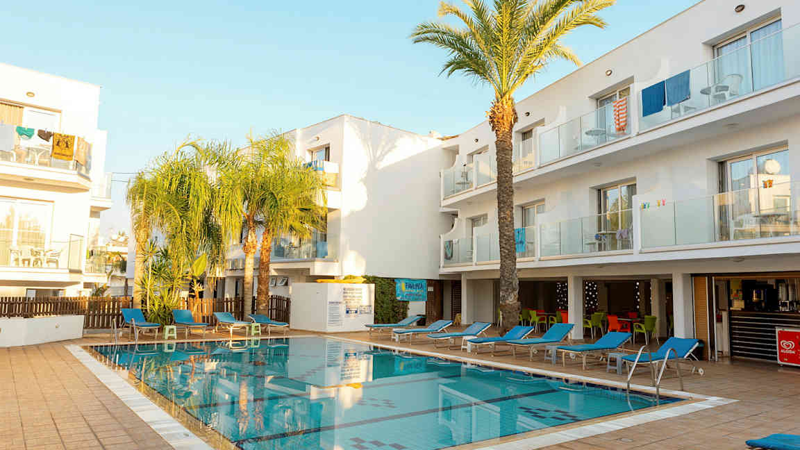 En utomhuspool omgiven av solstolar och vita byggnader. (AI-genererad text)