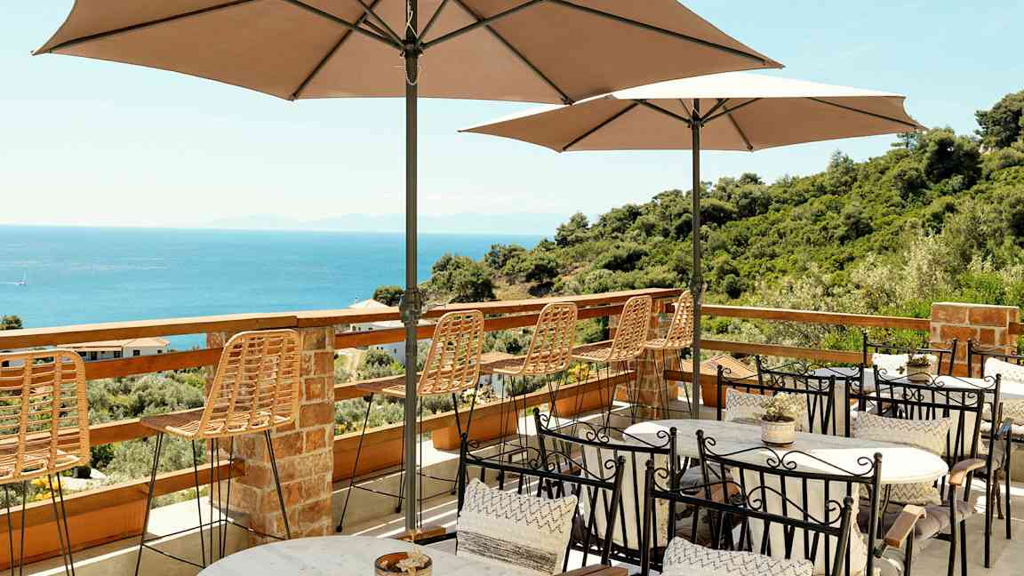 Terrass med bord, stolar och parasoller med utsikt över havet och grönskande omgivningar. (AI-genererad text)