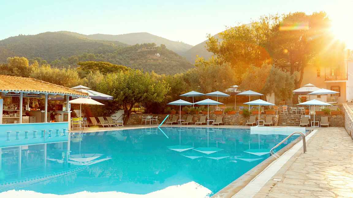 En utomhuspool med solstolar och parasoller, omgiven av träd och byggnader i ett bergigt landskap. (AI-genererad text)