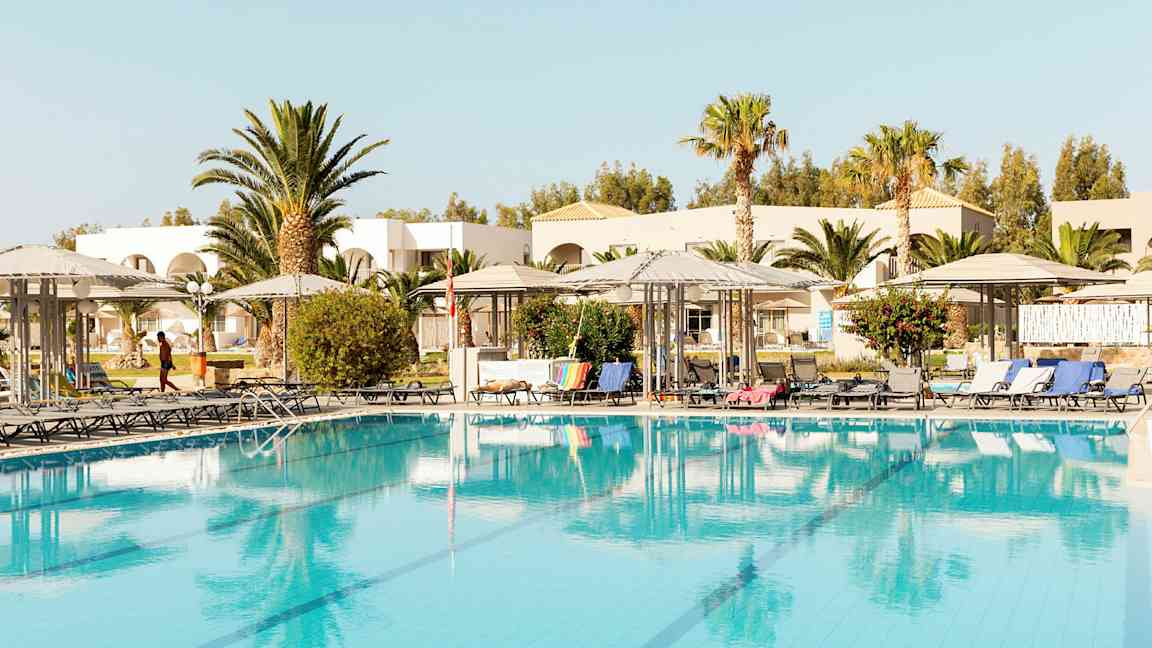 Bild av en pool med solsängar och parasoller, omgiven av träd och byggnader. (AI-genererad text)