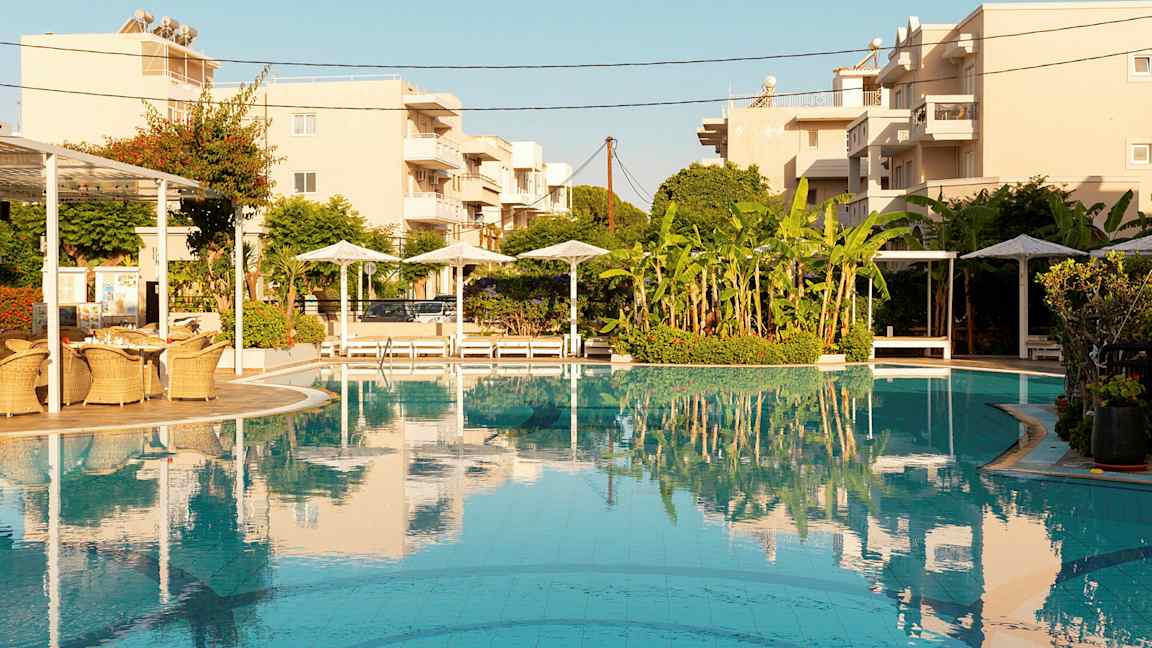 Poolområde med parasoller, solstolar och byggnader i bakgrunden. (AI-genererad text)