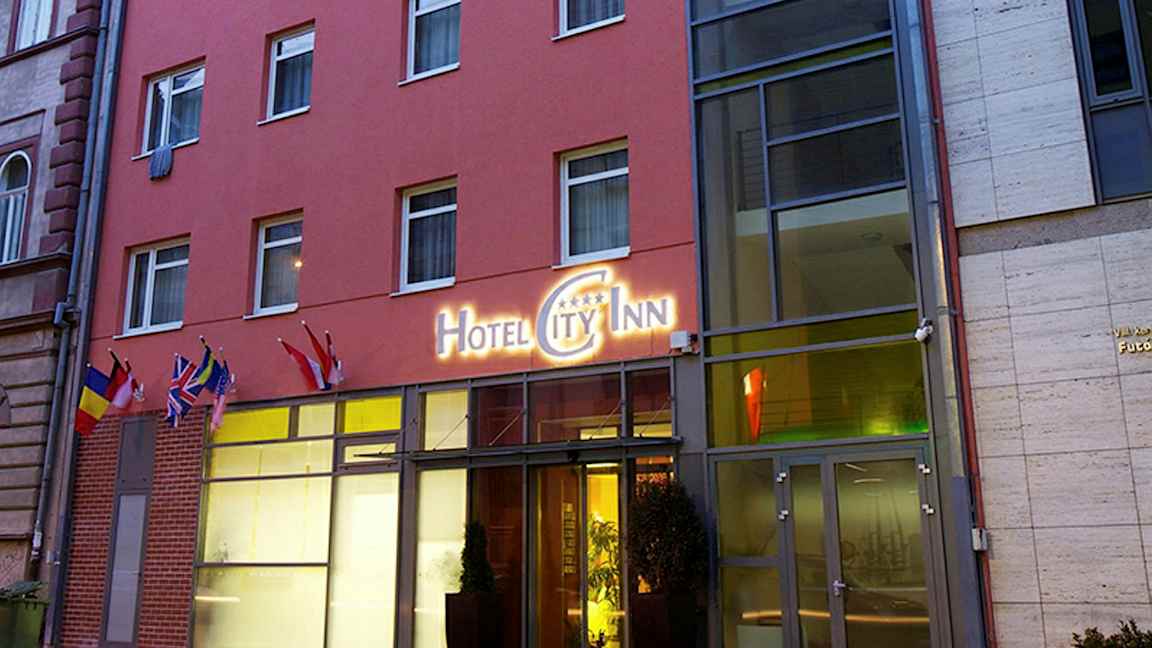 Utanför en hotellbyggnad med en skylt som lyder "Hotel City Inn" samt flera flaggor vid entrén. (AI-genererad text)