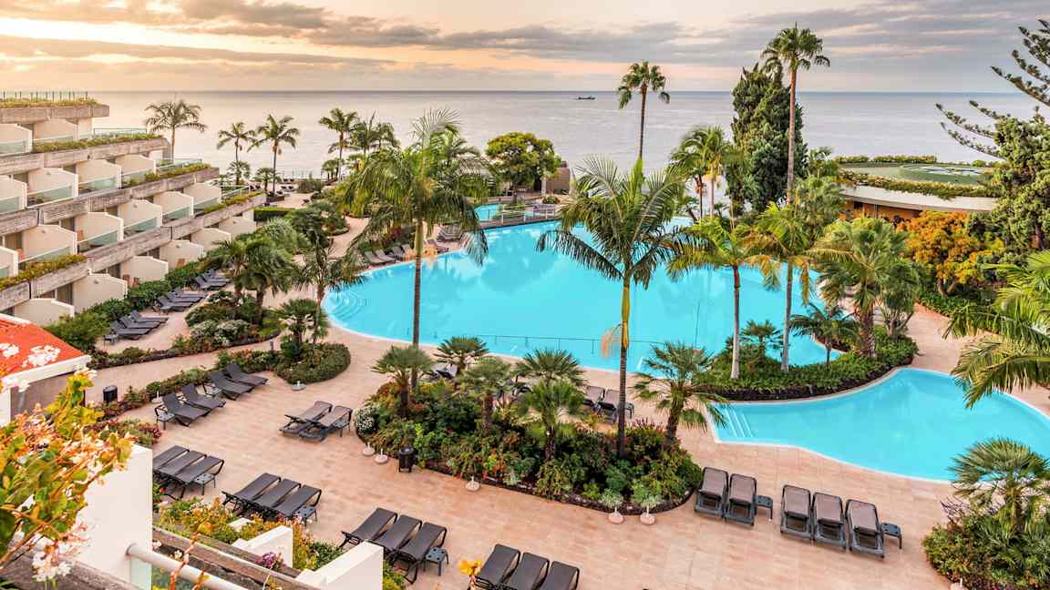 Bild på ett hotellområde med en stor swimmingpool omgiven av solstolar och tropiska växter. I bakgrunden syns havet och en solnedgång. (AI-genererad text)