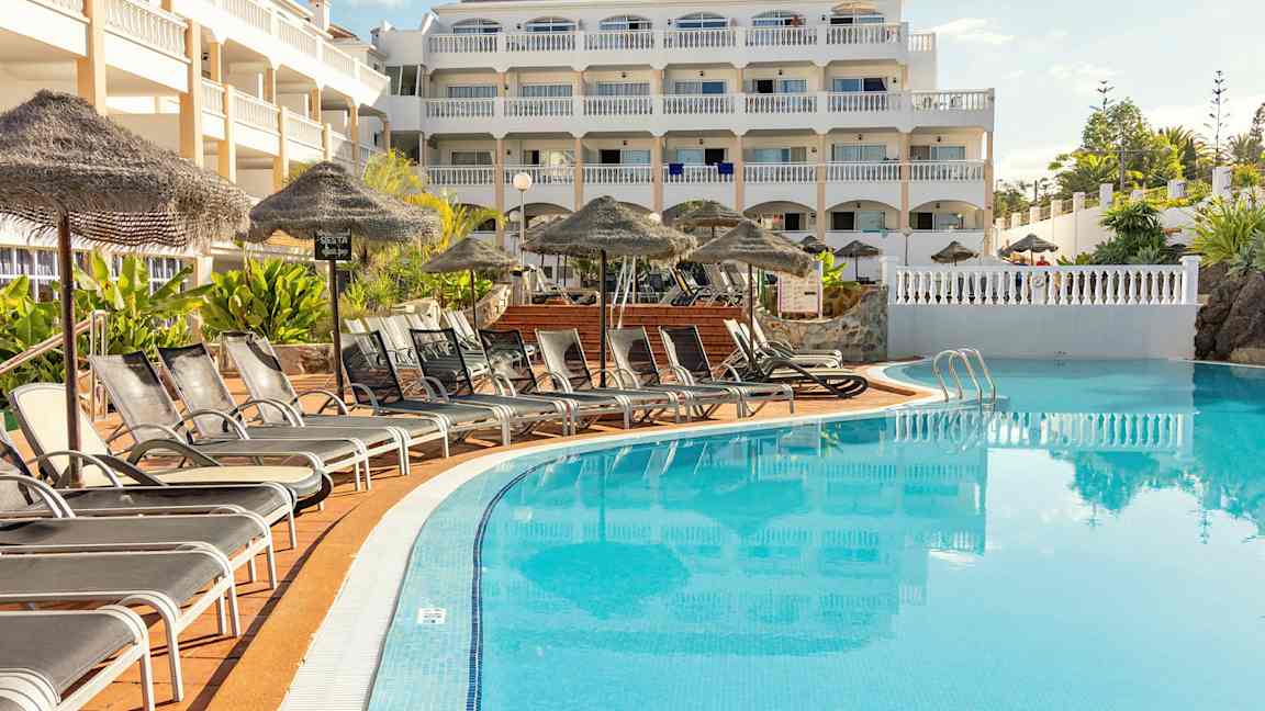 Bild av en hotellbyggnad med flera våningar som ligger vid en pool. Framför poolen står solstolar och parasoller. (AI-genererad text)