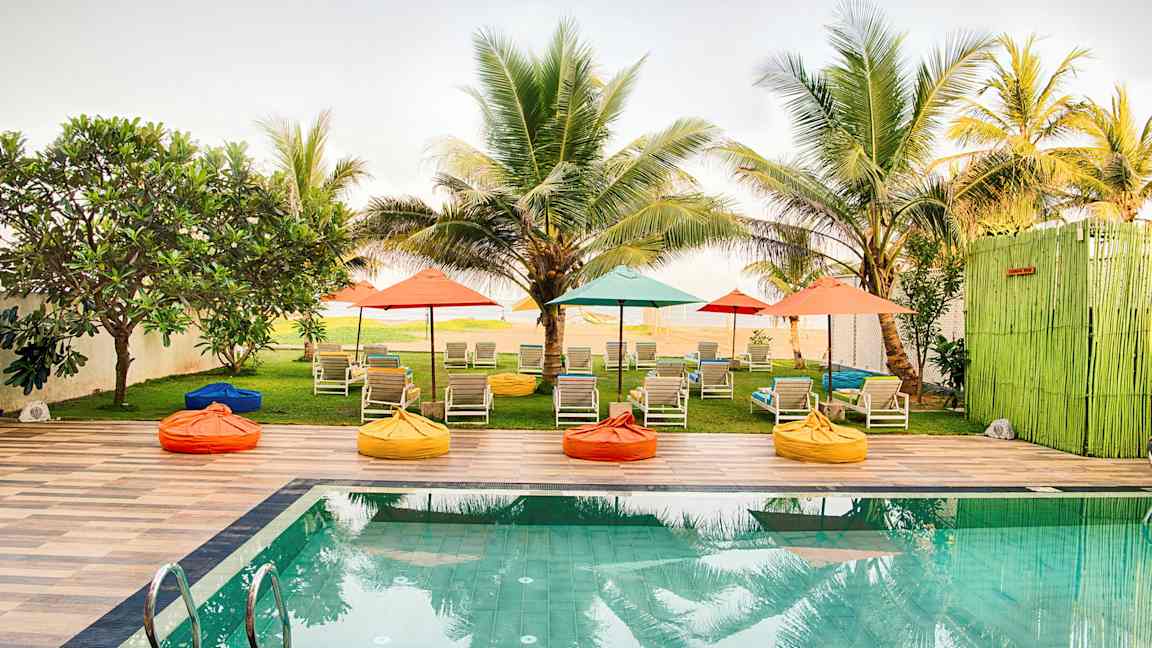 En pool med solsängar och färgglada parasoller i en trädgård omgiven av palmer. (AI-genererad text)
