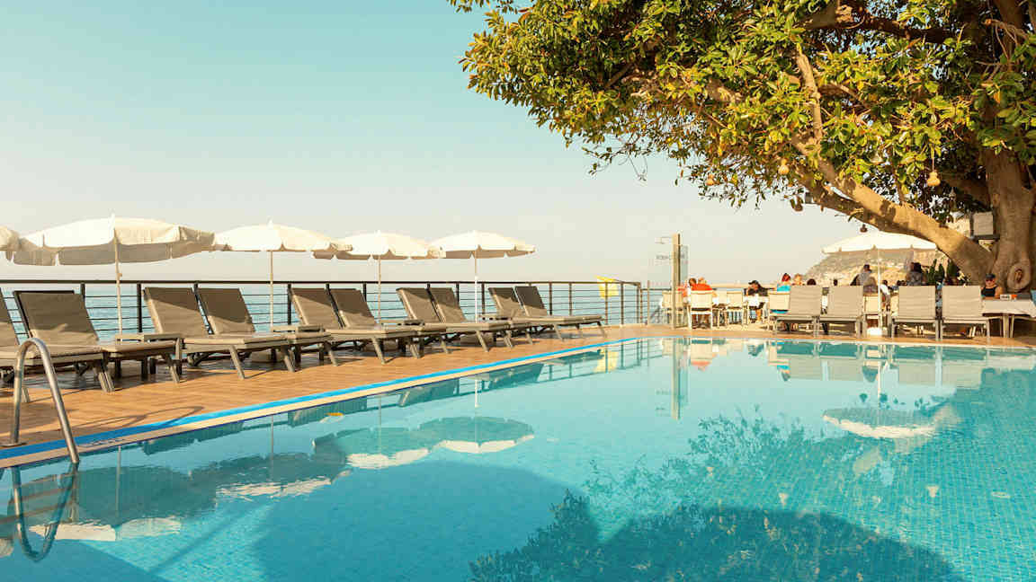 En utomhuspool med solstolar och parasoller bredvid, samt ett träd och ett trädäck med sittplatser. (AI-genererad text)