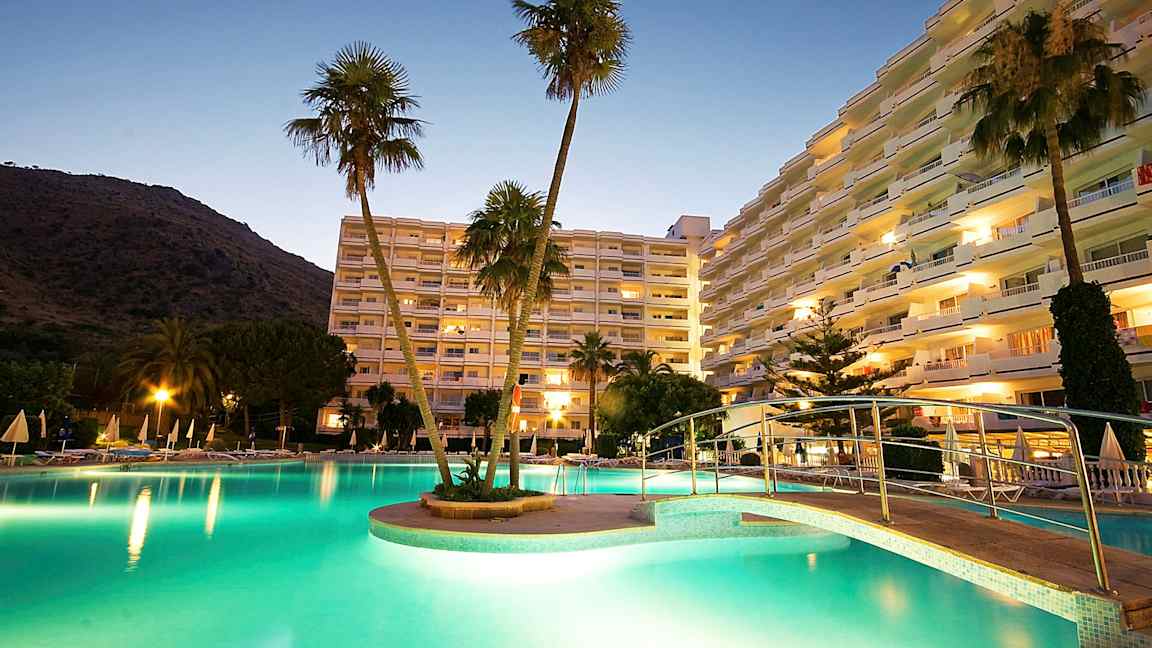 En bild på ett hotellområde vid skymning, med en upplyst pool, en bro och höga byggnader i bakgrunden. (AI-genererad text)
