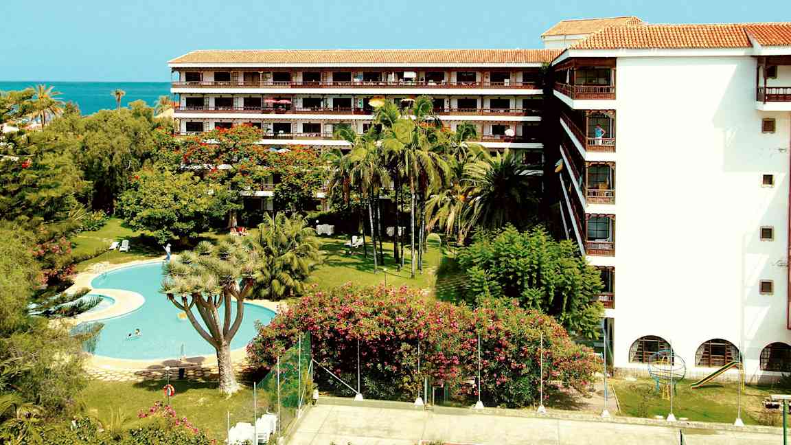 En hotellbyggnad med balkonger, omgiven av palmer och grönska, med en pool och en tennisbana i förgrunden. (AI-genererad text)