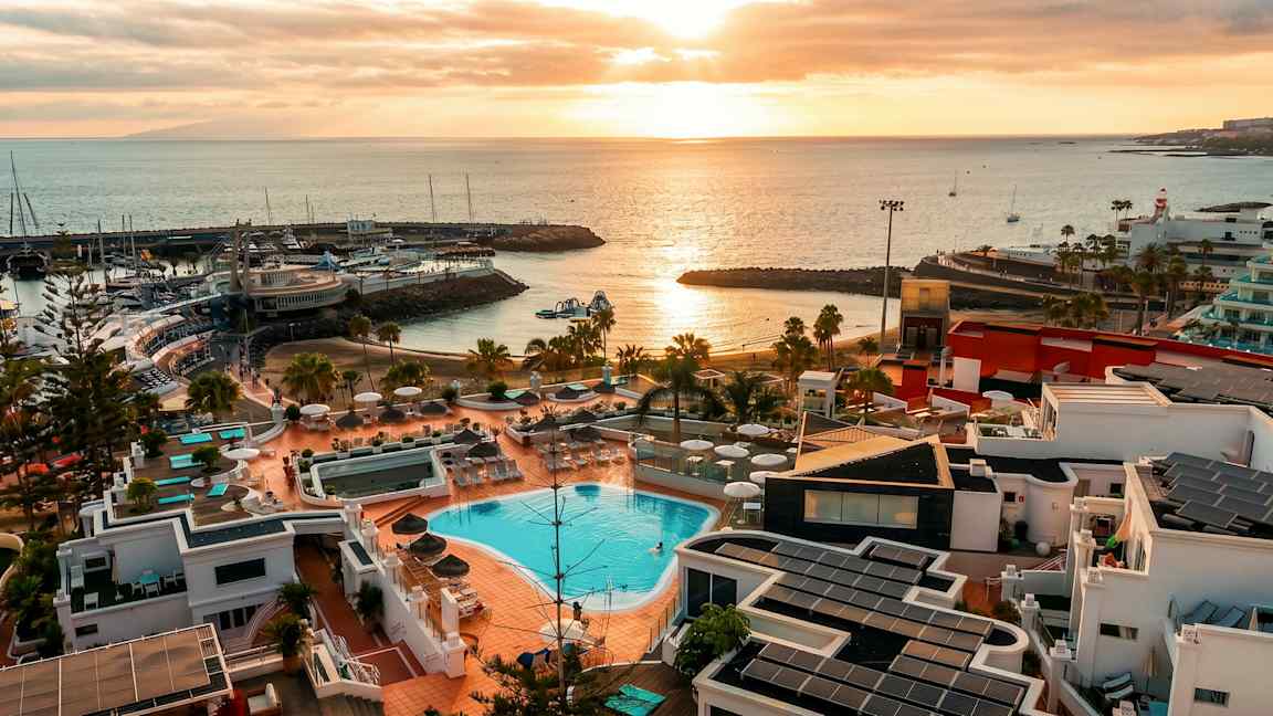 Bild av en kustöversikt med poolområde och byggnader i förgrunden, en hamn med båtar samt solnedgång över havet. (AI-genererad text)
