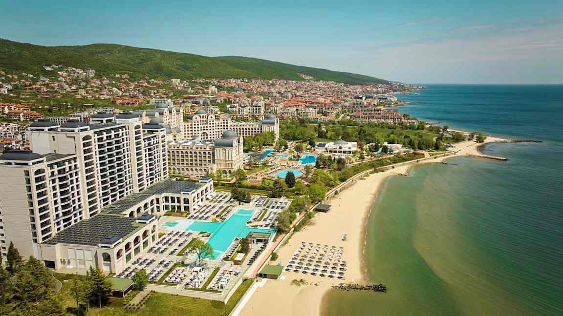 Hotell och pooler vid stranden med havet och staden i bakgrunden. (AI-genererad text)