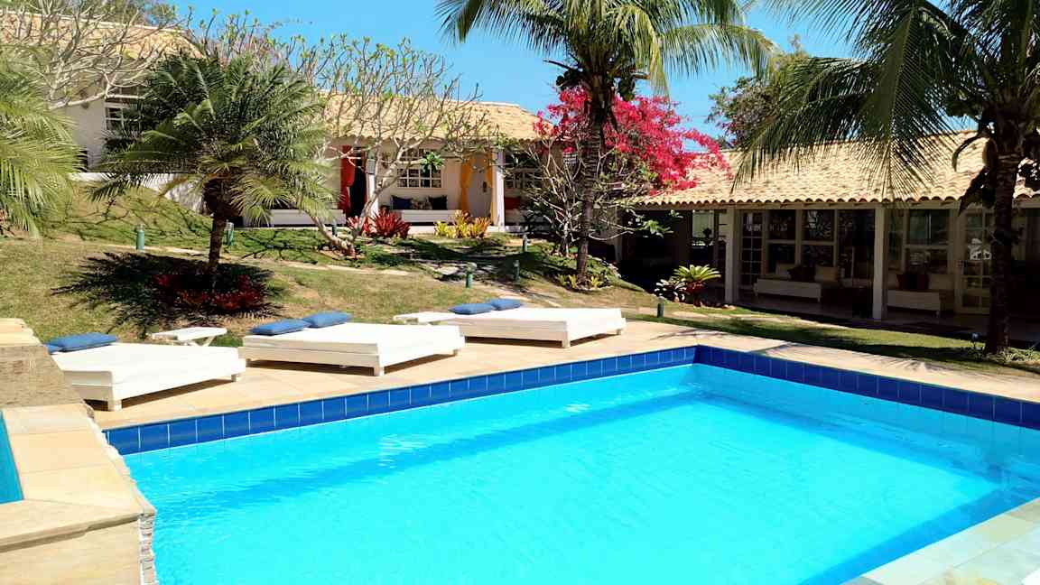 En pool omgiven av solstolar och tropiska växter vid en byggnad med terrakottatak. (AI-genererad text)