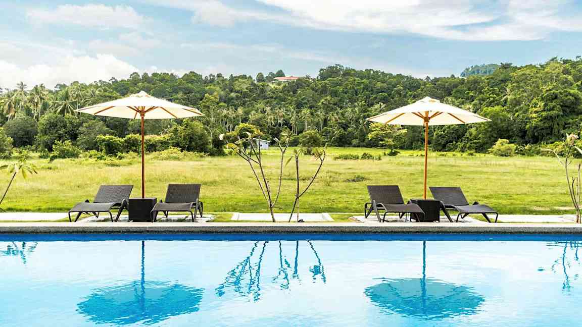 En pool med två solstolar och två vita parasoller framför ett grönskande fält. (AI-genererad text)