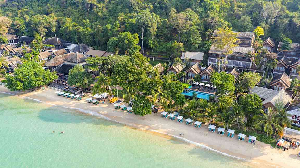 Flygfoto över en strand med solstolar och parasoller samt byggnader inbäddade i grönska. (AI-genererad text)