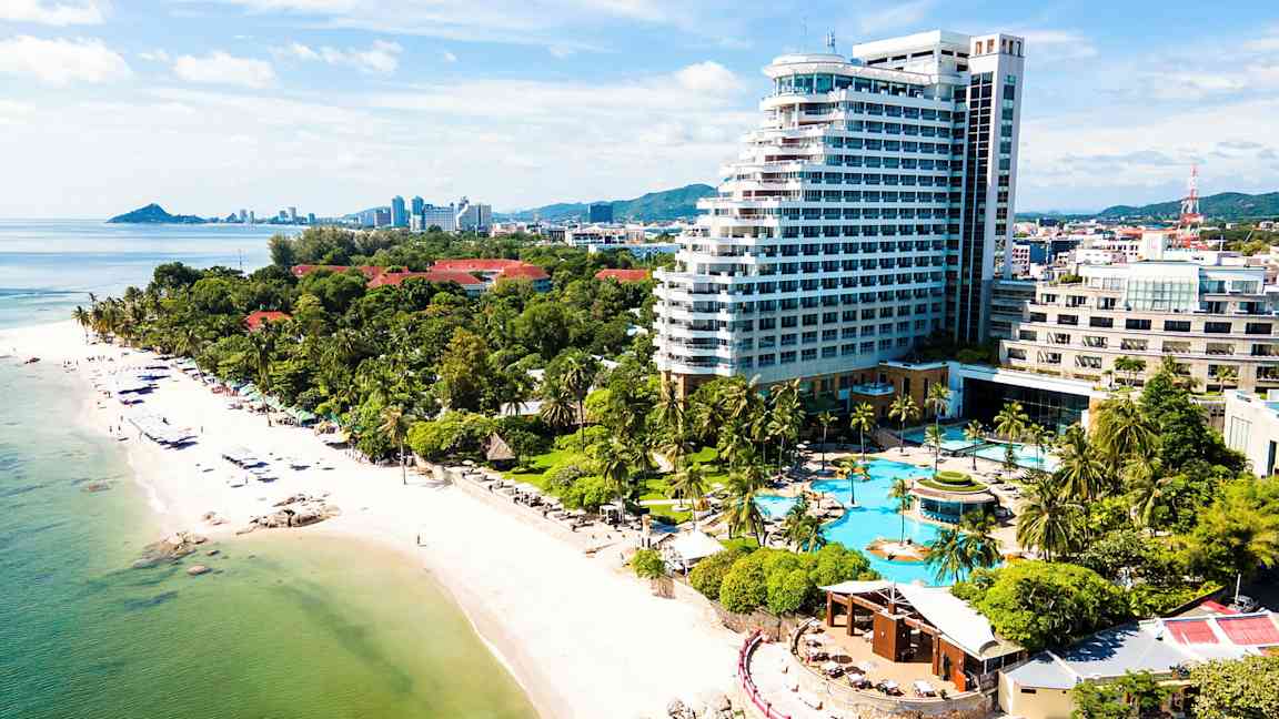 Bild av en kust med en sandstrand, palmer och ett stort hotellkomplex med poolområde. (AI-genererad text)