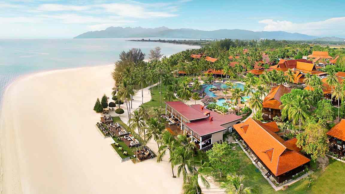 Strand med vit sand bredvid grönskande resort med orangea tak och poolområde. (AI-genererad text)