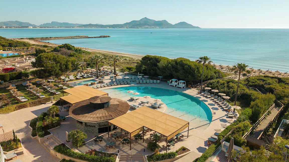 En swimmingpool omgiven av solstolar och parasoller, nära en strand och med utsikt över havet och berg i bakgrunden. (AI-genererad text)