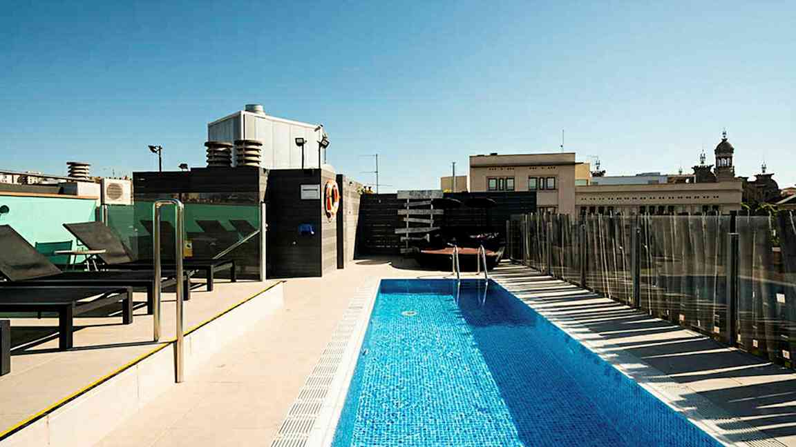 En utomhuspool på en takterrass omgiven av solstolar och byggnader i bakgrunden. (AI-genererad text)