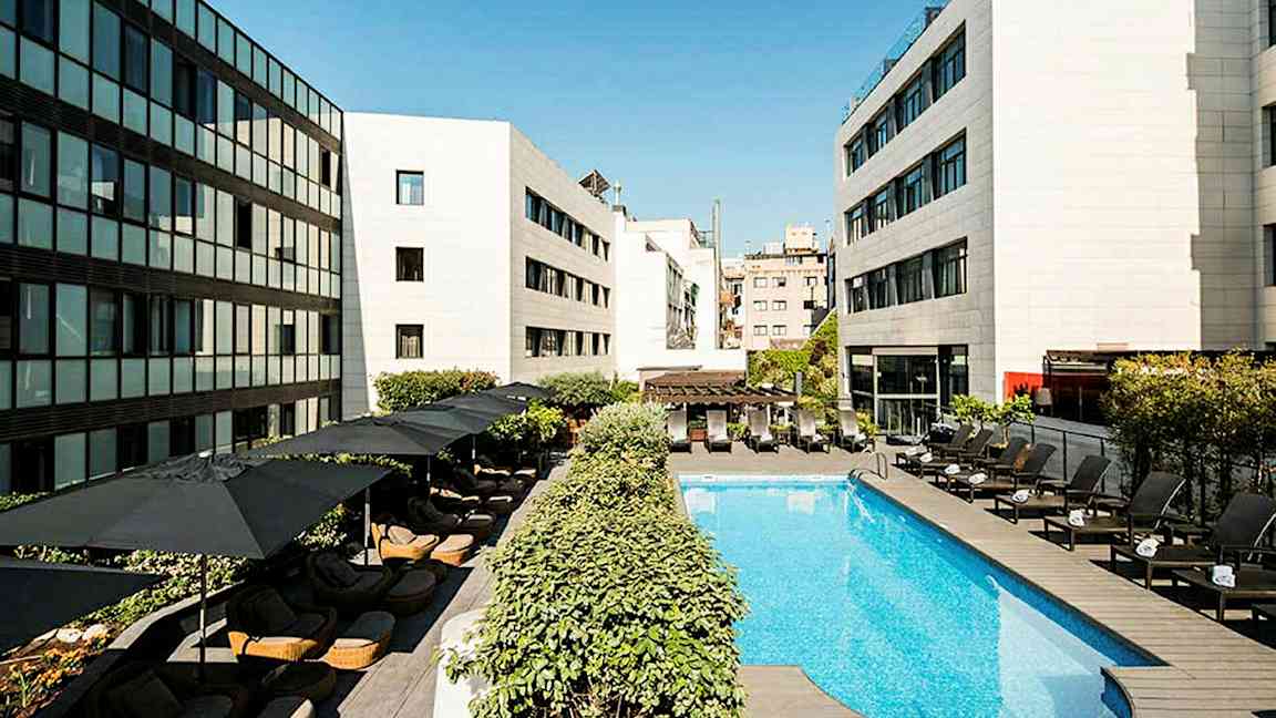 En hotellpool omgiven av liggstolar och parasoller, med moderna byggnader runtom. (AI-genererad text)