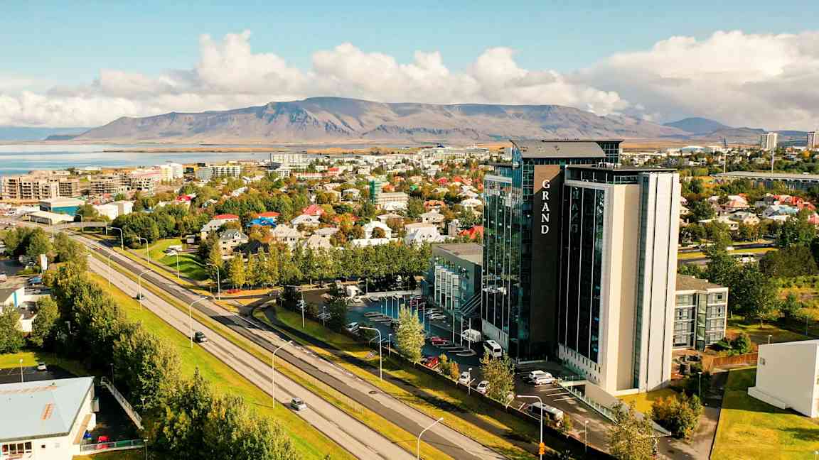 Vy över en stad med en stor byggnad märkt Grand i förgrunden, omgiven av bostadshus och grönska. I bakgrunden syns låga berg och en vattensamling. (AI-genererad text)