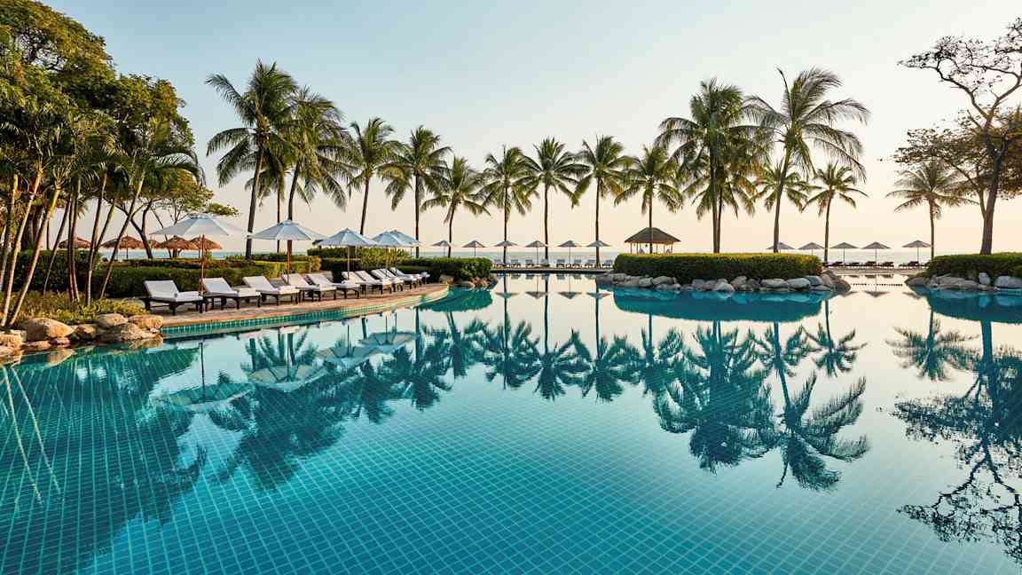 En utomhuspool med klarblått vatten, omgiven av solstolar, parasoller och palmer vid havet. (AI-genererad text)