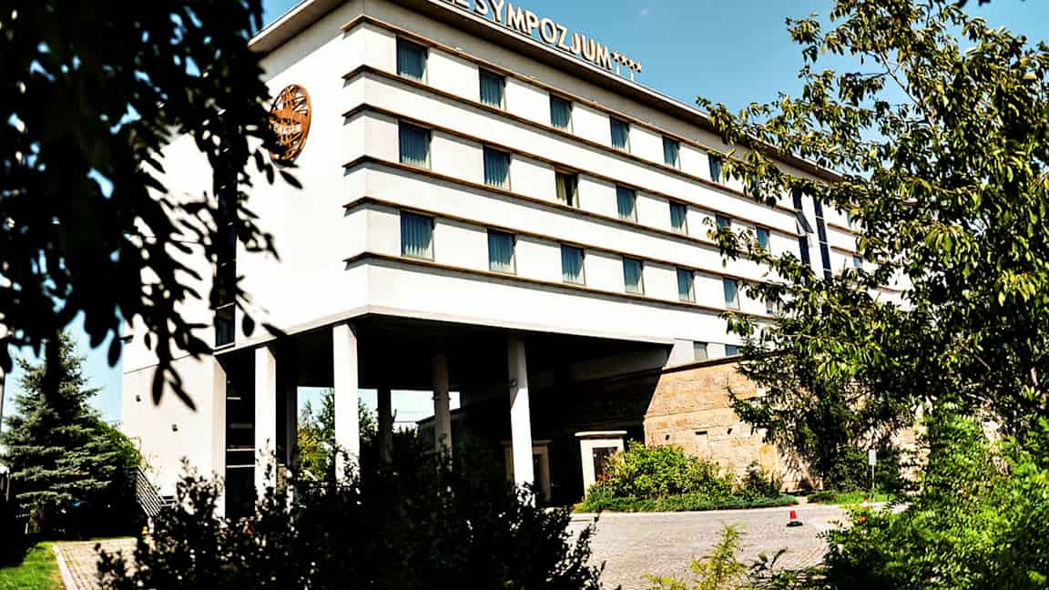 Byggnad med flera våningar, märkt med texten 'Hotel Sympozjum', omgiven av träd och växtlighet. (AI-genererad text)