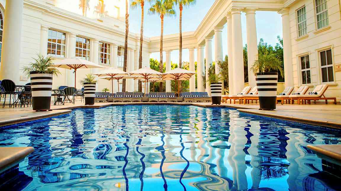 Poolområde med blanka blåa vatten, solstolar och parasoller omgivna av pelare och palmer. (AI-genererad text)