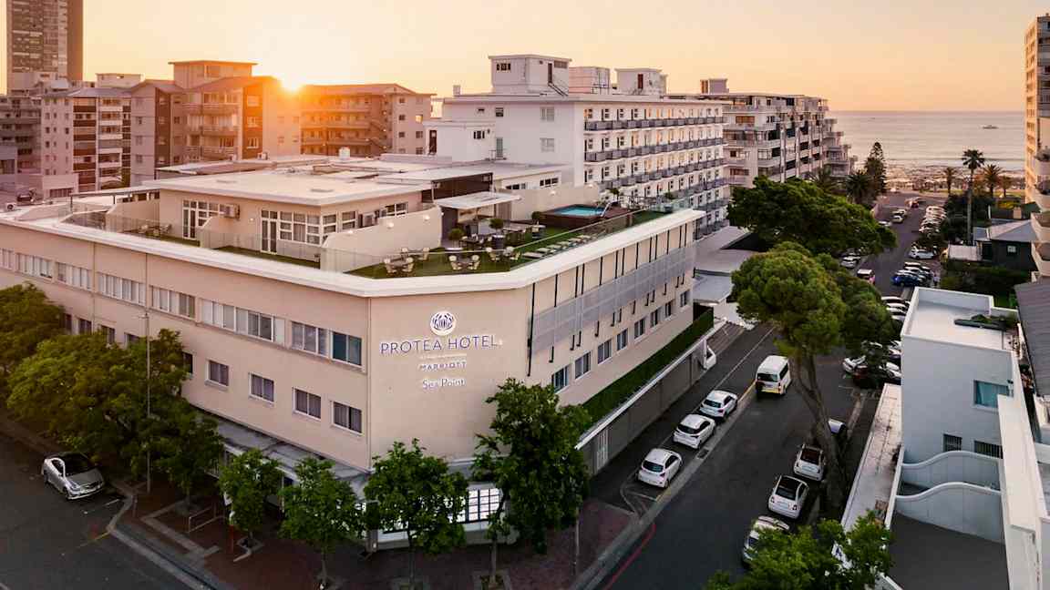 Hotellbyggnad vid havet omgiven av andra byggnader i solnedgång. (AI-genererad text)